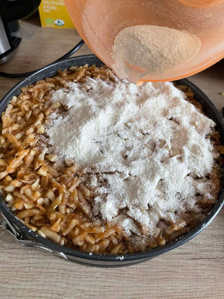 Obuolių pyragas su manais kepimo formoje, ant tarkuotų obuolių beriamas sausas mišinys