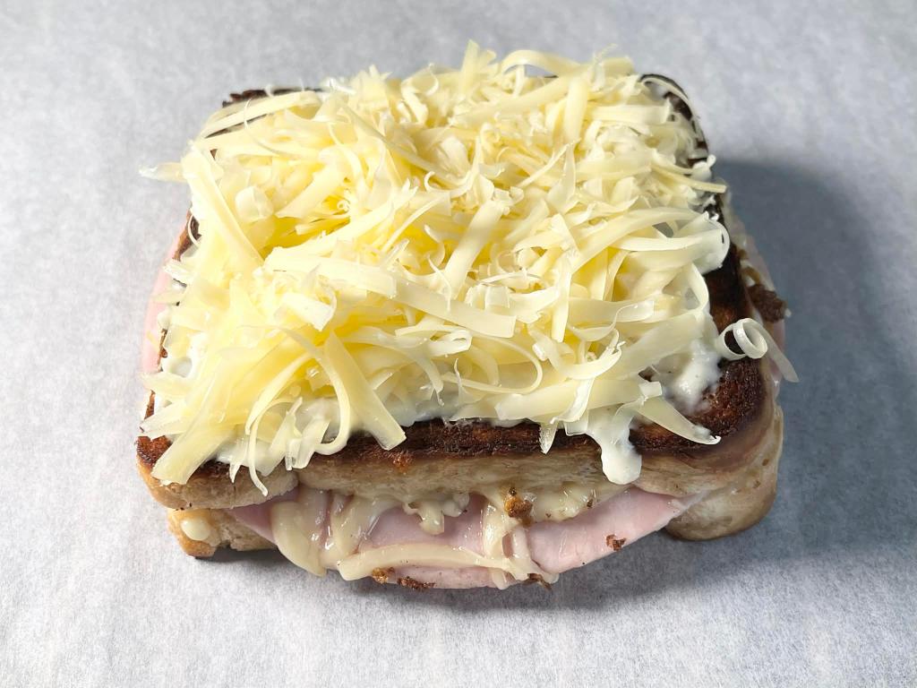 Apskrudintas Croque-monsieur karštas sumuštinis pateptas padažu ir apibarstytas tarkuotu sūriu