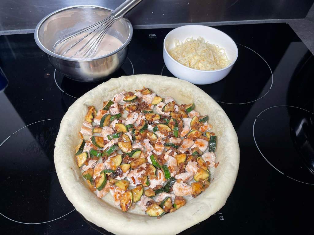 Ant kišo pagrindo apkepti ingredientai, dubenyje kiaušinių ir grietinėlės plakinys, indelyje tarkuotas sūris