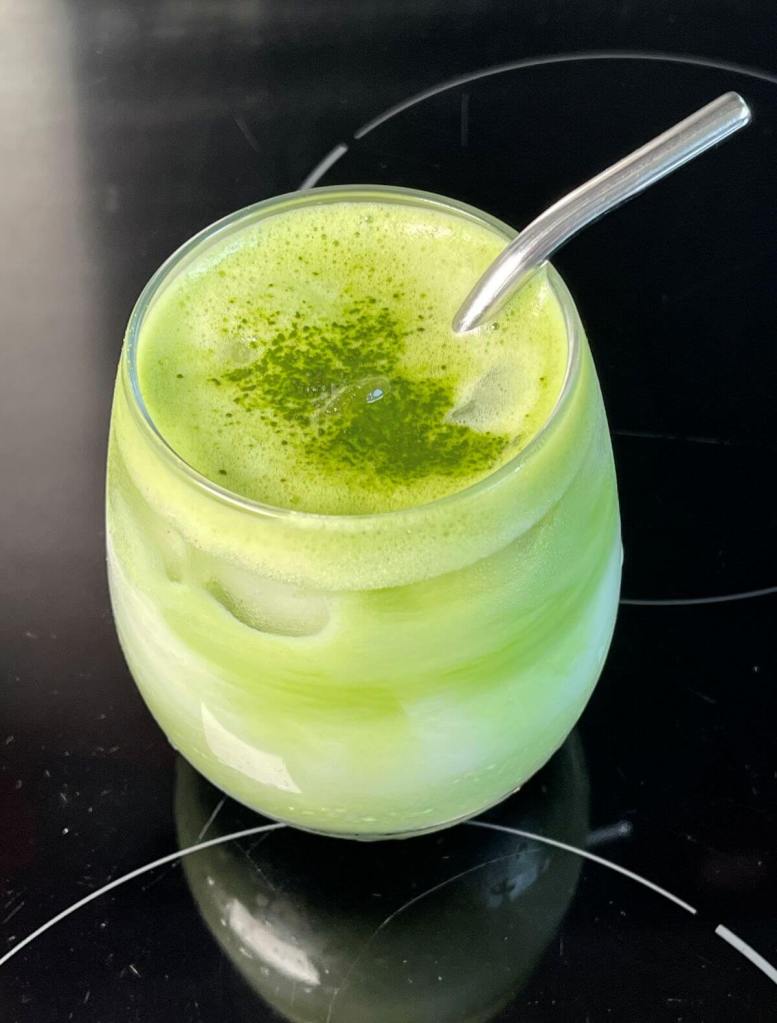 Ledinė matcha latte stiklinėje su daugkartiniu šiaudeliu ir pabarstyta matcha arbatos milteliais