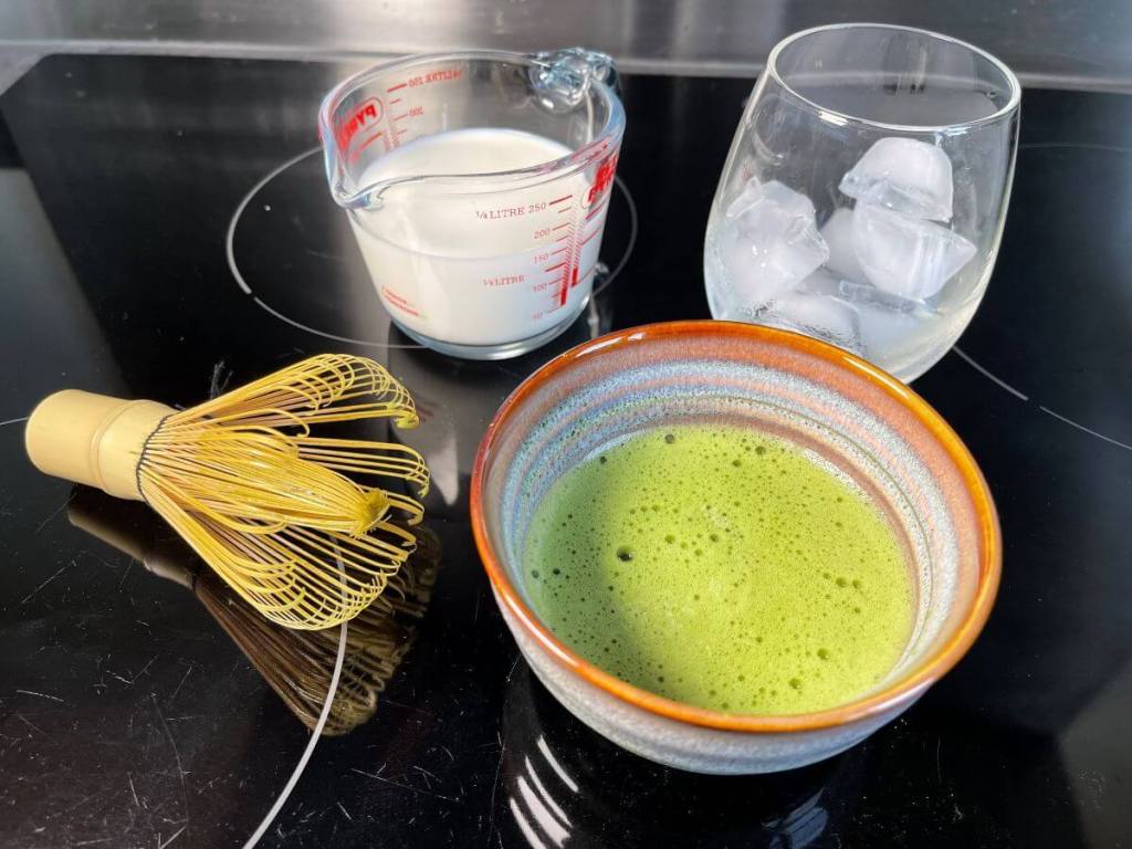 Dubenėlyje išplakta matcha su karštu vandeniu, pienas ir ledukai stiklinėje paruošti gaminti ledinę matcha latte