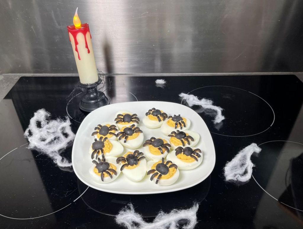 Velnio kiaušiniai įdaryti pikantišku įdaru su voriukais iš juodųjų alyvuogių, apibarstyti rūkyta paprika lėkštėje; Helovino dekoracijos: voratinkliai ir kruvina žvakė.